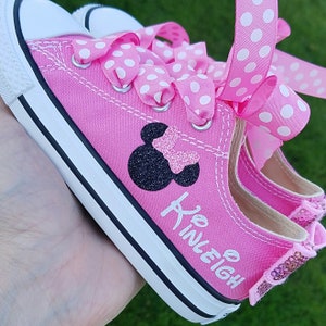 May include: Pink canvas children's shoes with black Mickey Mouse ears and a pink bow. The shoes have white polka dot ribbon laces and the name "Kinleigh" is written on the side.