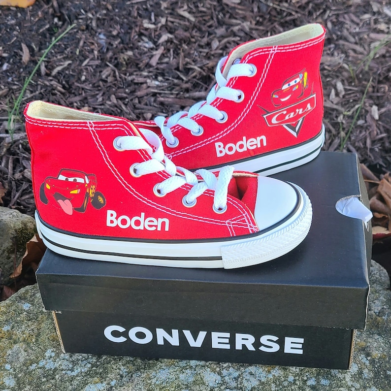 K&ouml;nnte beinhalten: Rote Converse-Sneaker mit hohem Schaft, einem Cars-Charakter-Motiv und dem Namen "Boden" auf jedem Schuh. Die Schuhe stehen auf einem schwarzen Converse-Schuhkarton.