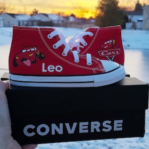 K&ouml;nnte beinhalten: Rote Converse High-Top Sneaker mit einem Lightning McQueen Cars-Charakterdesign. Die Schuhe haben wei&szlig;e Schn&uuml;rsenkel und eine wei&szlig;e Sohle. Der linke Schuh hat den Namen "Leo" an der Seite. Die Schuhe stehen auf einem schwarzen Converse Schuhkarton.
