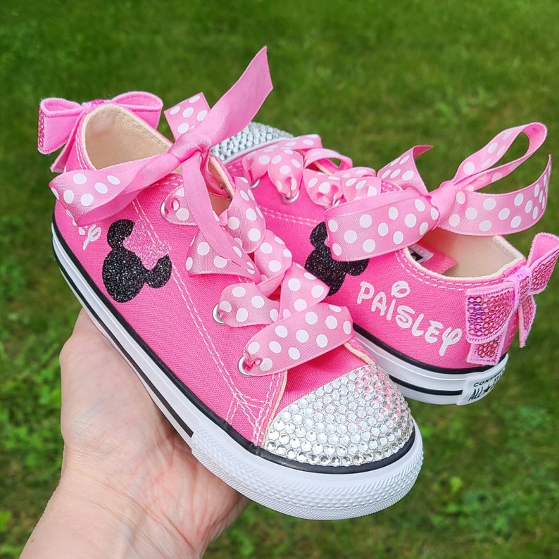 May include: Pink toddler-sized sneakers with white polka dot ribbon laces and a black glitter Minnie Mouse design. The shoes are decorated with rhinestones and the name "Paisley" is written on one shoe.