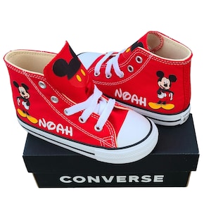 May include: Red Converse high-top sneakers featuring Mickey Mouse graphics and the name "NOAH" in white. The shoes have white laces, a white toe cap, and are displayed on a black Converse box.