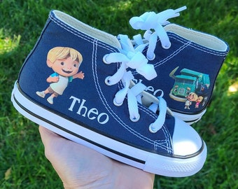 May include: Blue high-top canvas shoes with a cartoon character and the name "Theo" printed on the side. The shoes feature a white rubber sole and white laces.