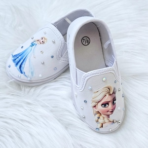 May include: White slip-on shoes featuring Elsa from Frozen. The shoes have a canvas upper, embellished with iridescent rhinestones. One shoe displays Elsa in a blue dress, while the other shows a close-up of her face. The size 7/8 is printed on the insole.