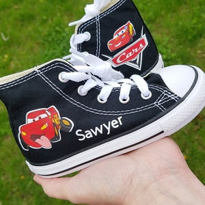 K&ouml;nnte beinhalten: Schwarze Canvas-High-Top-Sneaker mit wei&szlig;en Schn&uuml;rsenkeln und einem roten Comic-Auto-Motiv. Die Schuhe haben eine wei&szlig;e Gummisohle und der Text "Sawyer" und "Cars" ist auf den Schuhen aufgedruckt.