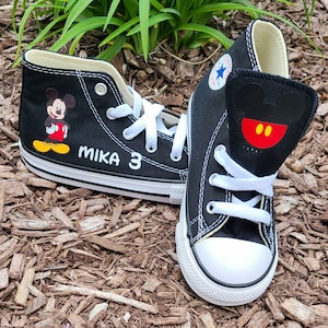 May include: Black high-top sneakers featuring Mickey Mouse graphics. One shoe displays Mickey's image, the other has a Mickey face design on the tongue. White laces and soles. The name "MIKA" and the number "3" are printed on one shoe.