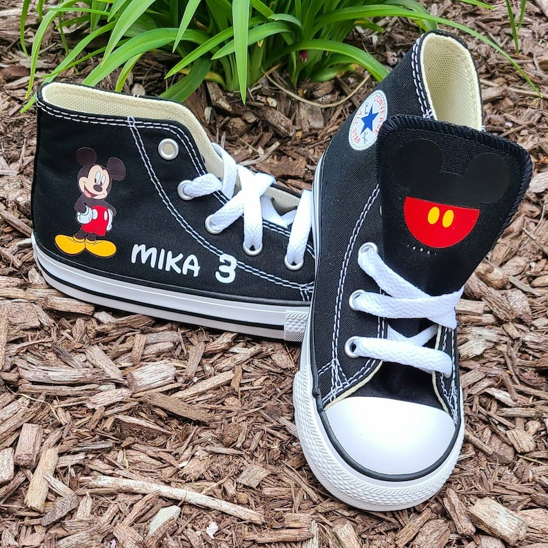 May include: Black high-top sneakers featuring Mickey Mouse graphics. One shoe displays Mickey's image, the other has a Mickey face design on the tongue. White laces and soles. The name "MIKA" and the number "3" are printed on one shoe.