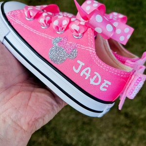 May include: Pink canvas sneakers with white polka dot ribbon laces and a silver glitter Minnie Mouse design. The name "JADE" is written in white on the side of the shoe.