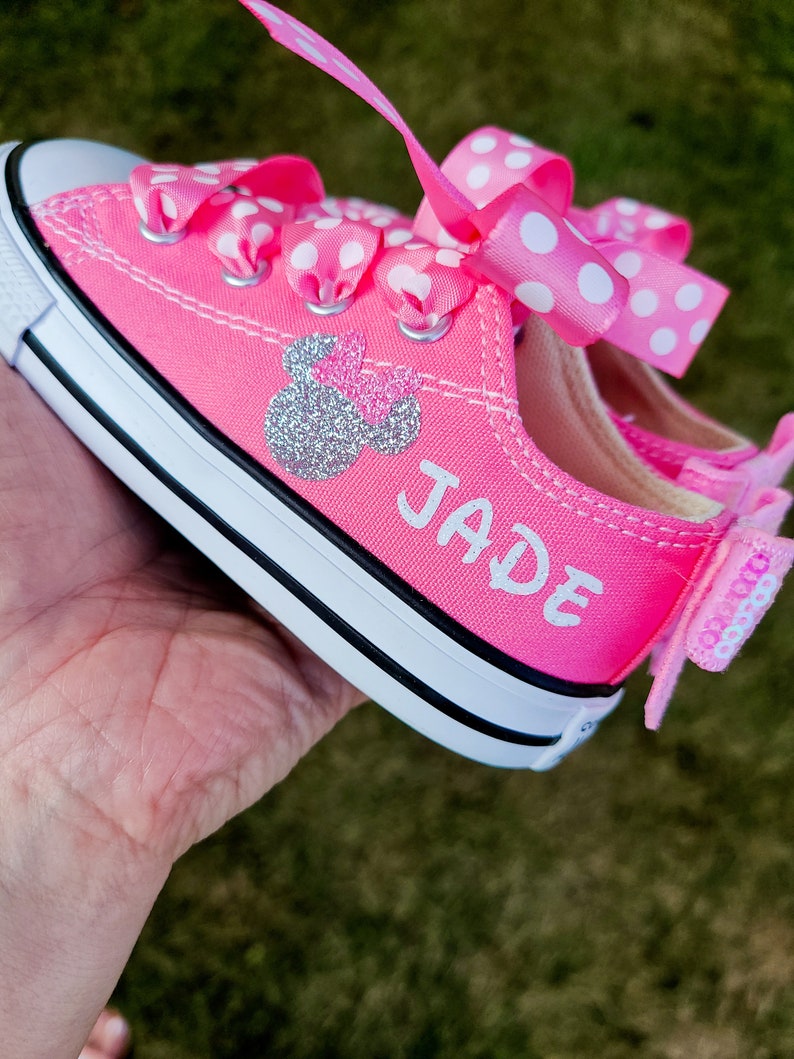 May include: Pink canvas sneakers with white polka dot ribbon laces and a silver glitter Minnie Mouse design. The name "JADE" is written in white on the side of the shoe.