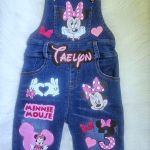 May include: Dark blue denim overalls featuring Minnie Mouse graphics, a pink glittery name patch reading CAELYN, a pink glittery number 3, and additional pink glittery heart and bow accents. The design includes white gloved hands forming a heart.