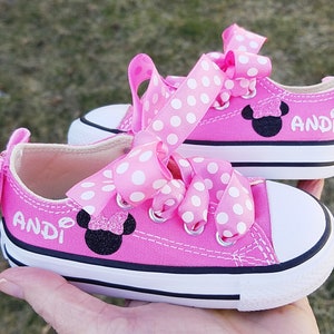 May include: Pink Converse sneakers with white polka dot ribbon laces and a black Minnie Mouse appliqu&eacute;. The shoes are personalized with the name "Andi".