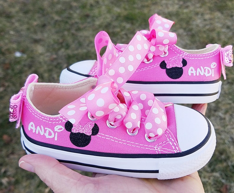 May include: Pink Converse sneakers with white polka dot ribbon laces and a black Minnie Mouse appliqu&eacute;. The shoes are personalized with the name "Andi".