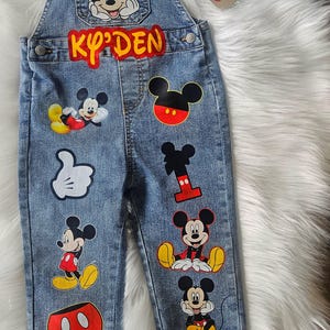 May include: Blue denim overalls for a child, adorned with various Mickey Mouse patches. The overalls feature the name "KY'DEN" in red and yellow lettering, along with multiple Mickey Mouse character designs.