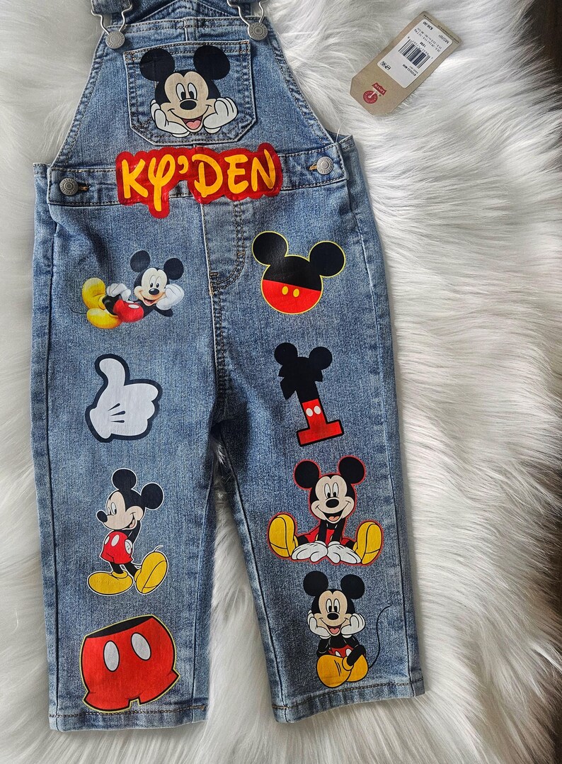 May include: Blue denim overalls for a child, adorned with various Mickey Mouse patches. The overalls feature the name "KY'DEN" in red and yellow lettering, along with multiple Mickey Mouse character designs.