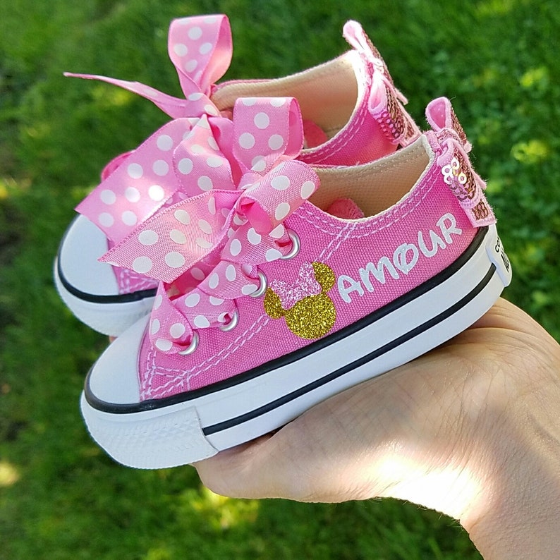May include: Pink baby shoes with white polka dots and a gold glitter Minnie Mouse design. The shoes have a white bow and the word "AMOUR" is written on the side.