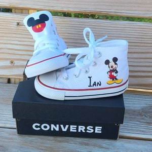 May include: White Converse high-top sneakers with red and blue accents, featuring a Mickey Mouse design and the name "Ian." The sneakers are displayed on a black Converse box. The shoes are designed for a child.