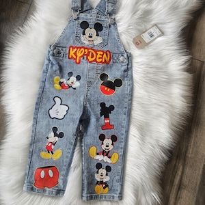 May include: Blue denim overalls featuring Mickey Mouse graphics. The overalls have a front pocket with a Mickey Mouse image and the name "KY'DEN" in red letters. Various Mickey Mouse designs are also on the legs.