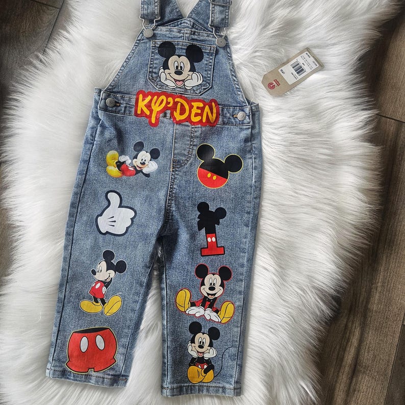 May include: Blue denim overalls featuring Mickey Mouse graphics. The overalls have a front pocket with a Mickey Mouse image and the name "KY'DEN" in red letters. Various Mickey Mouse designs are also on the legs.