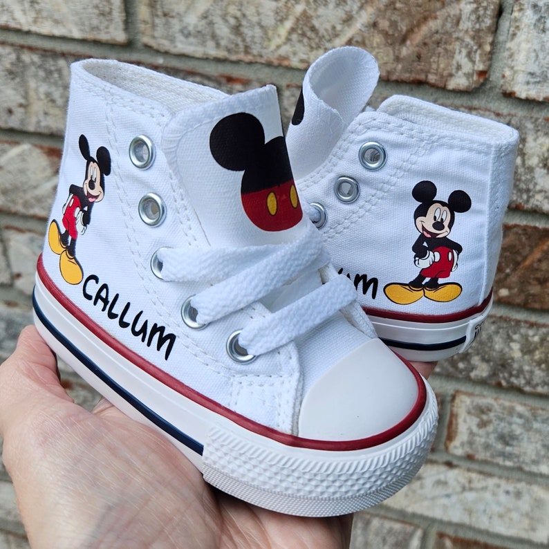 May include: White high-top canvas sneakers featuring a Mickey Mouse graphic on the sides. The shoes have red and blue accents on the soles and the name "Callum" printed on the side. The sneakers are designed for children.