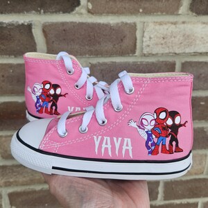 Puede incluir: Zapatillas altas de color rosa con suela blanca y un gr&aacute;fico de personajes de Spider-Man. Las zapatillas tienen el nombre "YAYA" escrito en el lateral.