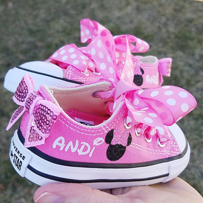 May include: Pink Converse sneakers with white polka dot bows and a black Mickey Mouse silhouette with a pink bow. The shoes are personalized with the name "Andi" in black.