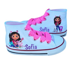 May include: White high-top sneakers with pink ribbon laces. The shoes feature a cartoon character illustration and the name "Sofia" in glitter. The soles have a red stripe.