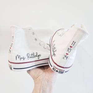 May include: White high-top Converse sneakers with the text "Mrs. Rutledge" and the date "11.10.24" written on them. The sneakers have red hearts next to the date.