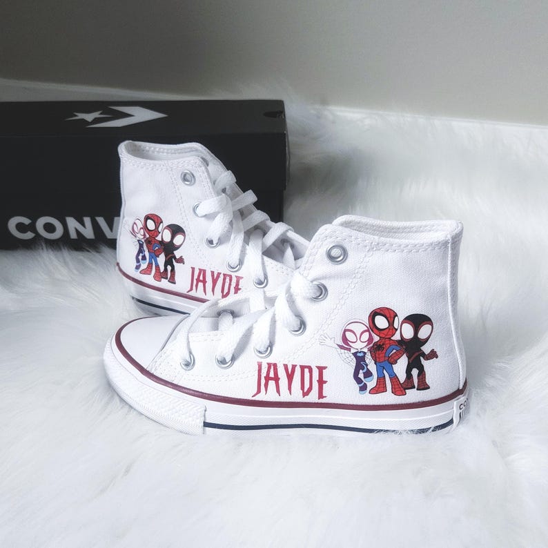 K&ouml;nnte beinhalten: Wei&szlig;e High-Top-Sneaker mit roten und blauen Akzenten, die ein Spider-Man-Motiv und den Namen "JAYDE" in Burgunderrot zeigen. Die Schuhe sind geschn&uuml;rt und stehen auf einer flauschigen wei&szlig;en Oberfl&auml;che, mit einer schwarzen Converse-Box im Hintergrund.