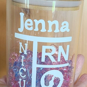 May include: A clear glass jar with a bamboo lid, filled with colorful beads. The jar is decorated with a white vinyl decal that says "Jenna RN" and includes a footprint symbol.