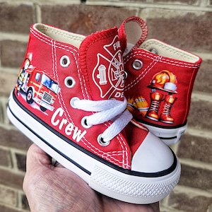 May include: Red high-top canvas sneakers with white laces and a white toe cap. The sneakers feature firefighter-themed graphics, including a fire truck, helmet, boots, and the word "Crew". The shoes have a black and white sole.