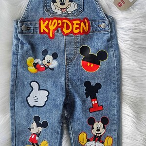 May include: Blue denim overalls for a child, adorned with various Mickey Mouse patches. The name "Ky'Den" is displayed in yellow and red lettering. Features multiple Mickey Mouse character designs.