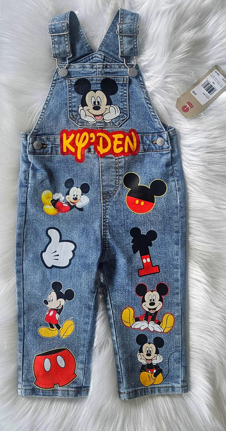 May include: Blue denim overalls for a child, adorned with various Mickey Mouse patches. The name "Ky'Den" is displayed in yellow and red lettering. Features multiple Mickey Mouse character designs.