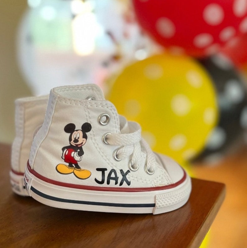 May include: White high-top baby sneakers with a Mickey Mouse graphic and the name "JAX" in black letters. The sneakers have red and blue accents on the sole and a white lace-up closure. Balloons are in the background.