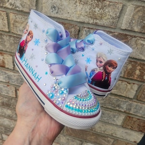 May include: White high-top canvas shoes with a sparkly design. The shoes feature a printed image of Elsa and Anna from Frozen, and the name "HANNAH" is written on the side. The shoes have blue and purple ribbon laces.
