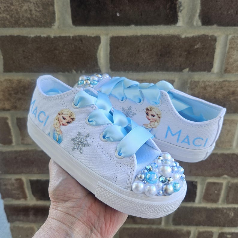 May include: White canvas sneakers featuring light blue satin ribbon laces, a cartoon character graphic, silver glitter snowflake accents, and the name MACI printed in blue. The rounded toe caps are decorated with blue and white pearls.