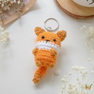 Puede incluir: Un llavero de zorro tejido a mano en naranja y blanco, con un anillo plateado. El zorro tiene pequeños ojos negros y una cola esponjosa. Un accesorio encantador.
