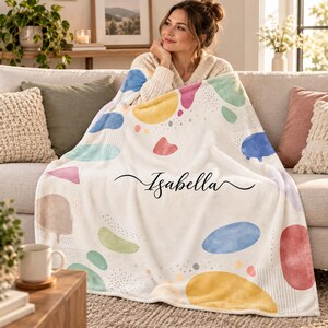 Op de afbeelding: Een zachte witte plaid met een kleurrijk patroon van abstracte vormen in blauw, geel, roze en groen, geaccentueerd met fijne lijntjes en stippen. De naam Isabella is in het midden gedrukt in een elegant, zwart cursief lettertype.