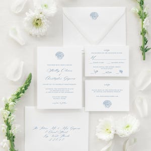May include: A sophisticated wedding invitation suite. White stationery with blue seashell motifs includes an invitation, RSVP card, and envelope. Floral and petal accents complement the design. The invitation text reads "Shelley Olivier & Christopher Gegnene".