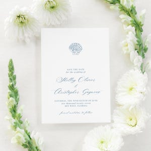May include: A white wedding invitation with blue text and a seashell graphic. The text reads "Save the Date" and lists the names Shelley Olivier and Christopher Gagnene, along with the wedding date and location.