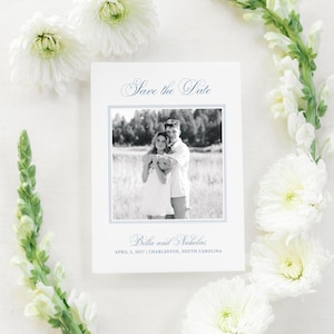 May include: A white Save the Date card with a blue border and text, featuring a black and white photo of a couple. The card is surrounded by white flowers and green stems. The text reads "Bella and Nicholas, April 3, 2027 | Charleston, South Carolina."