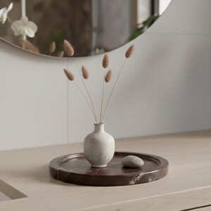 May include: A small, off-white ceramic vase holding dried, fluffy brown stems, set on a dark brown marble tray with a small, smooth stone. The tray sits on a light wood surface, with a round mirror in the background.