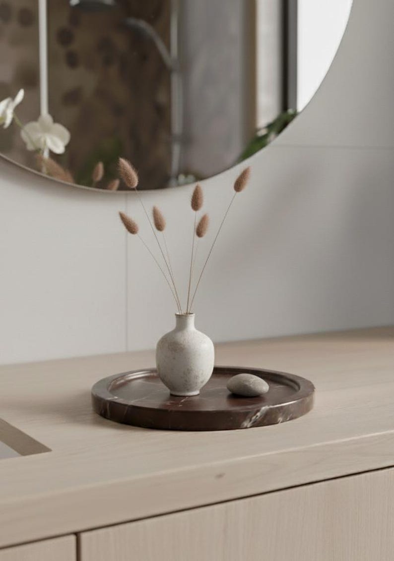 May include: A small, off-white ceramic vase holding dried, fluffy brown stems, set on a dark brown marble tray with a small, smooth stone. The tray sits on a light wood surface, with a round mirror in the background.