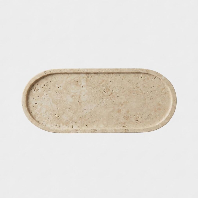 Pu&ograve; includere: Un vassoio di forma ovale realizzato in pietra travertino beige. La superficie presenta una consistenza naturale e porosa con varie venature color marrone chiaro e beige su tutto il materiale. Il vassoio ha un bordo rialzato.