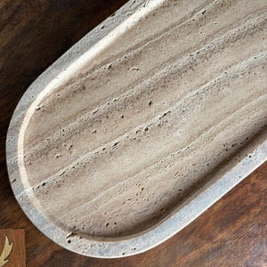 Pu&ograve; includere: Un vassoio di forma ovale realizzato in pietra travertino beige, caratterizzato da una texture naturale porosa e da distinti motivi di venature lineari sulla superficie. Il vassoio presenta un bordo rialzato liscio e una base poco profonda.