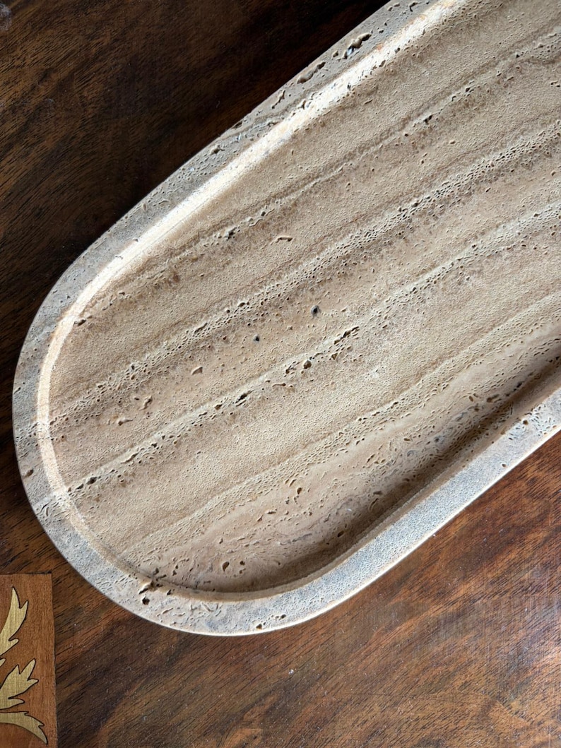 Pu&ograve; includere: Un vassoio di forma ovale realizzato in pietra travertino beige, caratterizzato da una texture naturale porosa e da distinti motivi di venature lineari sulla superficie. Il vassoio presenta un bordo rialzato liscio e una base poco profonda.