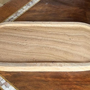 Pu&ograve; includere: Un vassoio di forma ovale realizzato in pietra travertino beige, caratterizzato da una texture naturale porosa e da distinti motivi a fasce orizzontali sulla superficie piana, con un bordo rialzato che circonda l'intero perimetro.