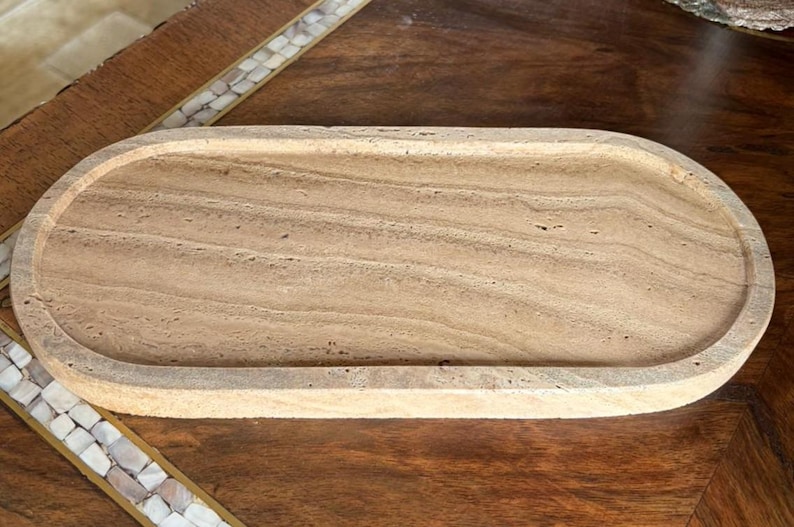 Pu&ograve; includere: Un vassoio di forma ovale realizzato in pietra travertino beige, caratterizzato da una texture naturale porosa e da distinti motivi a fasce orizzontali sulla superficie piana, con un bordo rialzato che circonda l'intero perimetro.