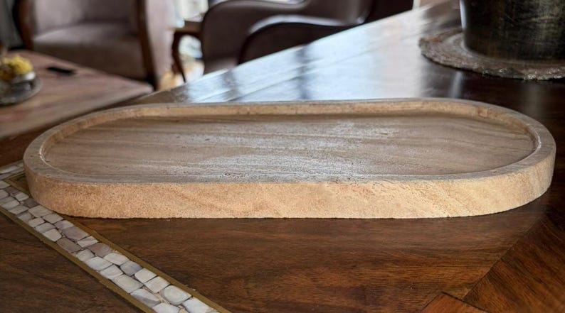 Pu&ograve; includere: Un vassoio in legno di forma ovale con una finitura color marrone chiaro e venature naturali del legno. Il vassoio presenta un centro poco profondo e un bordo liscio e arrotondato, offrendo un design semplice per riporre piccoli oggetti.