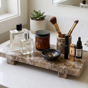 May include: A rectangular brown marble riser tray holding two glass perfume bottles, a brown glass jar with a black lid, a marble brush holder with two makeup brushes, a small black bowl with rings, a black mascara tube, a gold lipstick, and a dropper bottle.