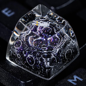 May include: A clear resin mechanical keyboard keycap featuring a detailed, textured lunar surface in deep black and purple tones. A tiny white astronaut figure with yellow accents is positioned on the cratered surface, encased within the transparent top.