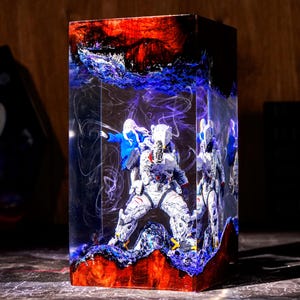 May include: A rectangular resin block containing a miniature astronaut figure in a white suit and a figure in a blue jacket. The resin features swirling white smoke-like patterns, deep red sections at the top and bottom, and blue accents.